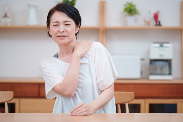 テーブルで肩を押さえてつらそうな更年期の女性